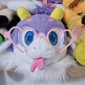 Puede incluir: Una cabeza de animal de peluche morada y blanca con gafas de corazón rosa y una lengua rosa que sobresale.