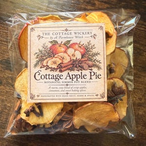 Op de afbeelding: Een doorzichtige plastic zak met een botanische sudderpotmix met het label The Cottage Wickery by A Farmhouse Witch Cottage Apple Pie. De mix bevat gedroogde appelschijfjes, kaneelstokjes, steranijs en hele kruidnagels.