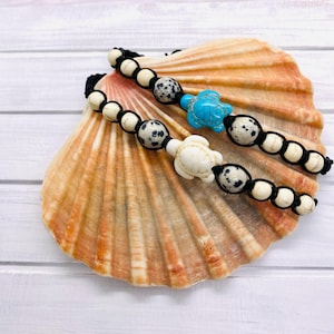 Op de afbeelding: Twee armbanden met een zwart koord en witte en bruine gevlekte kralen. Elke armband heeft een schildpadbedel, een is blauw en de andere is wit. De armbanden worden gepresenteerd op een schelp.