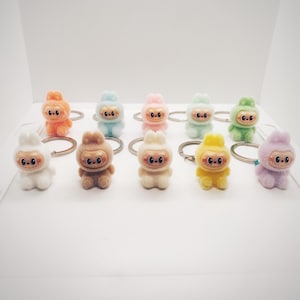 May include: A collection of ten colorful plush keychains in various pastel shades, including orange, blue, pink, green, yellow, and purple. Each keychain features a small, stylized figure with a round face and a silver keyring.