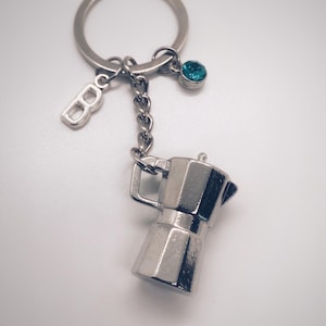 Puede incluir: Llavero plateado con un dije de cafetera italiana en miniatura. El llavero incluye un pequeño dije con la letra "B" y una gema de color turquesa. El dije de la cafetera es detallado y brillante.