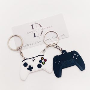 Peut inclure: Un ensemble de deux porte-clés représentant une manette de jeu vidéo blanche et une noire. Les porte-clés sont attachés à des anneaux de clés argentés. Le porte-clés blanc a un petit charme argenté avec la lettre "A" dessus. La carte en arrière-plan indique "Lovely Jewels, Merci de nous avoir choisis."