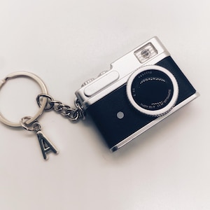 Op de afbeelding: Een zwart en zilveren camera sleutelhanger met een zilveren ketting en sleutelring. De sleutelhanger heeft een kleine zilveren letter "A" bedel. De camera heeft een lens en flitser.