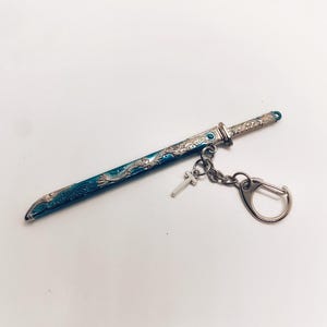 Peut inclure: Miniature porte-clés en forme d'épée décorative avec un design bleu sarcelle et argenté. L'épée présente des détails complexes et un petit charme en forme de croix. Le porte-clés comprend un fermoir argenté.