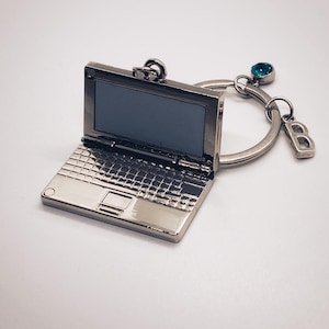 Può includere: Portachiavi a forma di laptop argentato con un ciondolo di gemma blu e un ciondolo con la lettera "B". Il laptop si apre per rivelare uno spazio vuoto per una foto.
