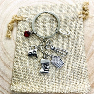 Op de afbeelding: Een zilveren sleutelhanger met een rode edelsteenbedel en verschillende bakthema-bedels, waaronder een maatbeker, garde, cupcake en een standmixer. De sleutelhanger zit in een klein jutezakje.