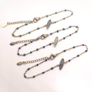 Op de afbeelding: Vier gouden kettingarmbanden met kleine surfplank bedels. De bedels zijn versierd met blauw, groen en rood email. Twee armbanden hebben blauwe kralen en twee hebben groene kralen.