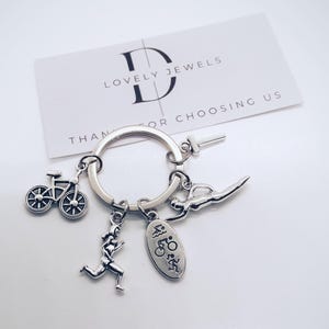 Puede incluir: Llavero de plata con dijes que representan los deportes de triatlón: una bicicleta, un corredor, un nadador y un dije con la palabra "Triatlón".