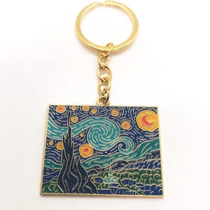 Könnte beinhalten: Ein goldfarbener Schlüsselanhänger mit einem quadratischen Emaille-Anhänger, der Vincent van Goghs Gemälde "Sternennacht" darstellt. Der Anhänger hat einen blauen Hintergrund mit gelben Sternen, einem wirbelnden Himmel und einer Sichelmond.