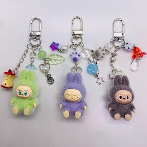 Op de afbeelding: Drie pluche konijnensleutelhangers in groen, paars en grijs. Elke sleutelhanger heeft een zilveren sluiting, decoratieve kralen en een kleine konijnenfiguur met een unieke gezichtsuitdrukking. De sleutelhangers hebben verschillende bedels, waaronder sterren, harten en een kleine appel.