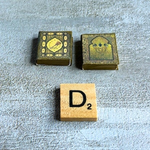 May include: Two miniature, ornate books with gold and green designs, and a wooden Scrabble tile with the letter "D" and the number "2". The books have detailed patterns and are placed on a textured, light gray surface.