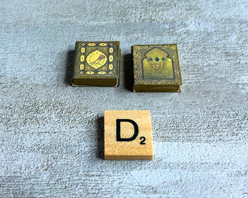 May include: Two miniature, ornate books with gold and green designs, and a wooden Scrabble tile with the letter "D" and the number "2". The books have detailed patterns and are placed on a textured, light gray surface.