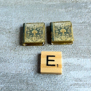 May include: Two miniature books with teal and gold covers, featuring a tree design, sit beside a wooden Scrabble tile with the letter "E" and the number "1". The books have gold-colored edges.