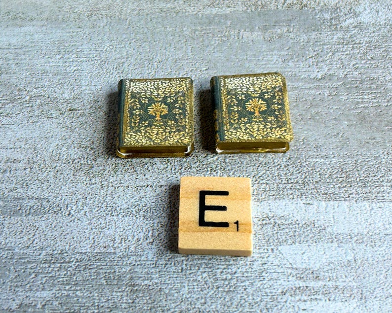 May include: Two miniature books with teal and gold covers, featuring a tree design, sit beside a wooden Scrabble tile with the letter "E" and the number "1". The books have gold-colored edges.