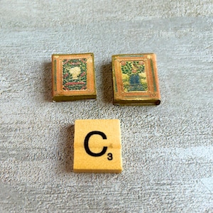 May include: Two miniature vintage books with ornate covers and a Scrabble tile with the letter "C" and the number "3" on a light gray wooden surface. The books have detailed illustrations and a gold border.