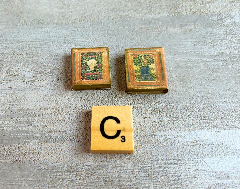 May include: Two miniature vintage books with ornate covers and a Scrabble tile with the letter "C" and the number "3" on a light gray wooden surface. The books have detailed illustrations and a gold border.