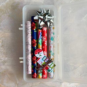 May include: A clear plastic storage container filled with rolled wrapping paper, a silver bow, scissors, and gift tags. The wrapping paper rolls feature various patterns, including floral, snowflakes, and geometric designs. A red tag reads "Holiday Ribbon".