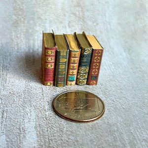 May include: A collection of five miniature books with gold-colored pages and decorative covers in red, green, and gold. A coin is in the foreground. The books are standing upright, showcasing their spines.
