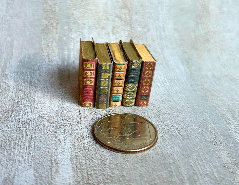 May include: A collection of five miniature books with gold-colored pages and decorative covers in red, green, and gold. A coin is in the foreground. The books are standing upright, showcasing their spines.