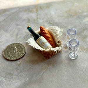 May include: A miniature wine bottle, two wine glasses, and a loaf of bread in a wicker basket. The bottle has a green label with the word "Beaujolais" on it. The glasses are clear and the bread is brown.