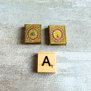 May include: Two small, ornate, square boxes with gold and green floral designs and a wooden tile with the letter "A" and the number "1". The boxes have intricate patterns with red, pink, and blue accents. The tile is a light beige color.