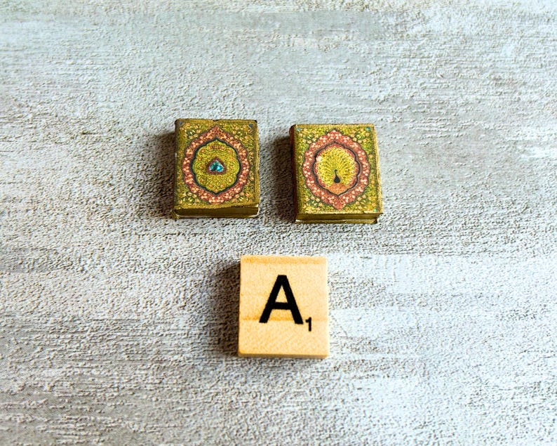 May include: Two small, ornate, square boxes with gold and green floral designs and a wooden tile with the letter "A" and the number "1". The boxes have intricate patterns with red, pink, and blue accents. The tile is a light beige color.
