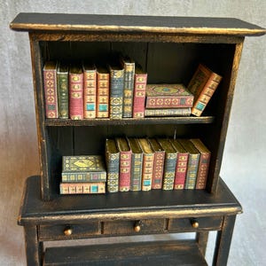 May include: A miniature black bookshelf with gold trim, filled with small, decorative books. The books have colorful spines and intricate designs, creating a vintage aesthetic. The bookshelf has two shelves and a small desk with two drawers.