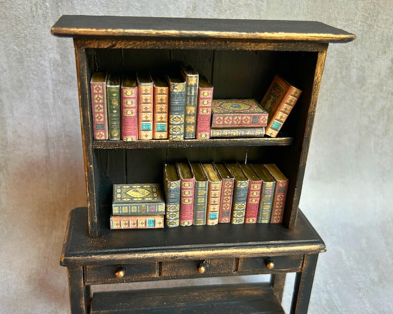 May include: A miniature black bookshelf with gold trim, filled with small, decorative books. The books have colorful spines and intricate designs, creating a vintage aesthetic. The bookshelf has two shelves and a small desk with two drawers.