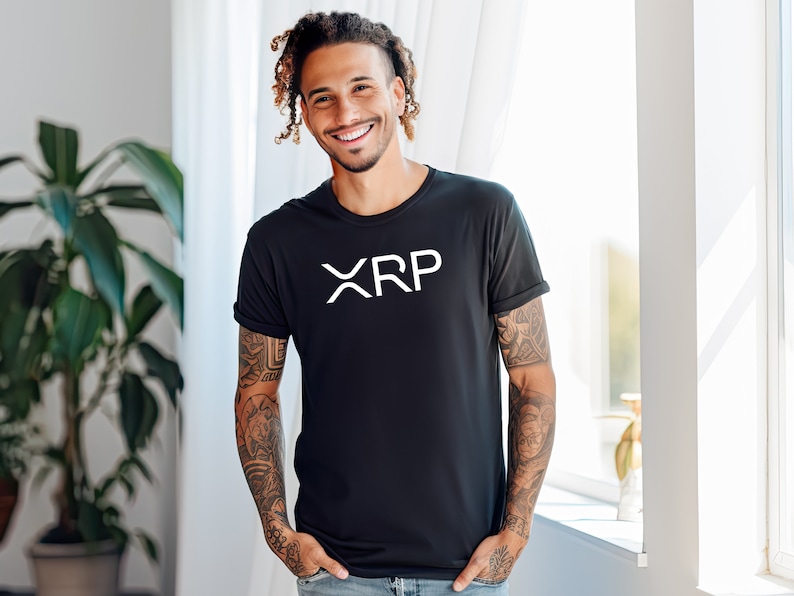 May include: A man with tattoos and dreadlocks is wearing a black t-shirt with the word "XRP" printed in white on the front. He is smiling and looking at the camera.