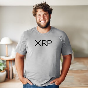 May include: A man wearing a gray t-shirt with the word "XRP" printed in black on the front. He is standing in a room with a wooden floor and a lamp on a side table.