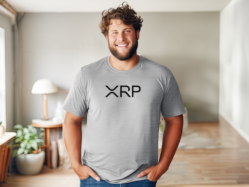 May include: A man wearing a gray t-shirt with the word "XRP" printed in black on the front. He is standing in a room with a wooden floor and a lamp on a side table.