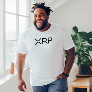 May include: A man wearing a white t-shirt with the word "XRP" printed in black on the front. He is smiling and standing in front of a window.