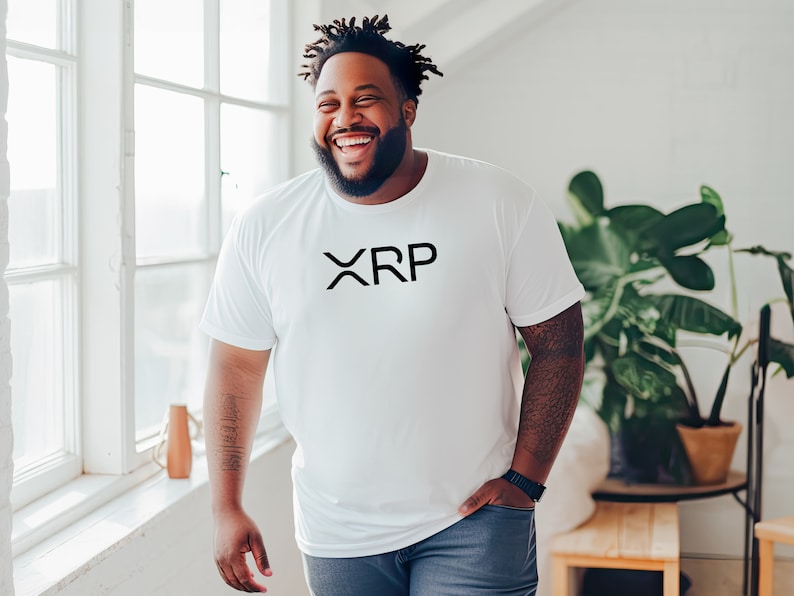 May include: A man wearing a white t-shirt with the word "XRP" printed in black on the front. He is smiling and standing in front of a window.