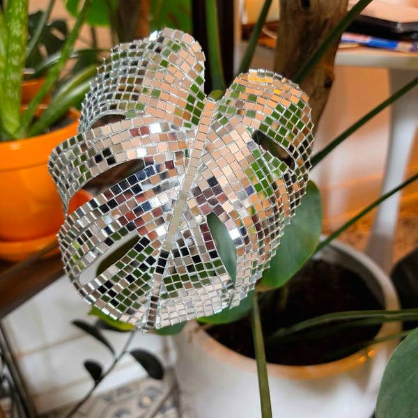 May include: A decorative leaf sculpture, covered in small, reflective mirror tiles, sits among green plants. The leaf is a Monstera shape, with its characteristic holes. The sculpture adds a touch of sparkle and a unique decorative element to the plant.