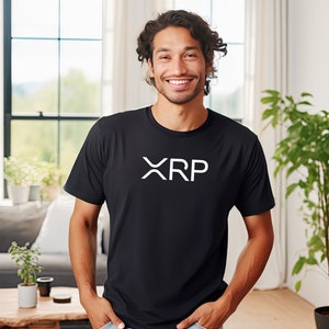 May include: A man wearing a black t-shirt with a white "XRP" logo. He is smiling and standing in a room with a window and plants.