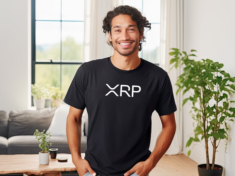 May include: A man wearing a black t-shirt with a white "XRP" logo. He is smiling and standing in a room with a window and plants.