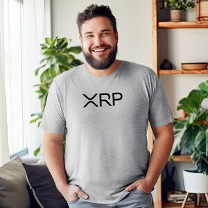 May include: A man wearing a gray t-shirt with the word "XRP" printed in black on the front. He is smiling and standing in a room with a window and plants.