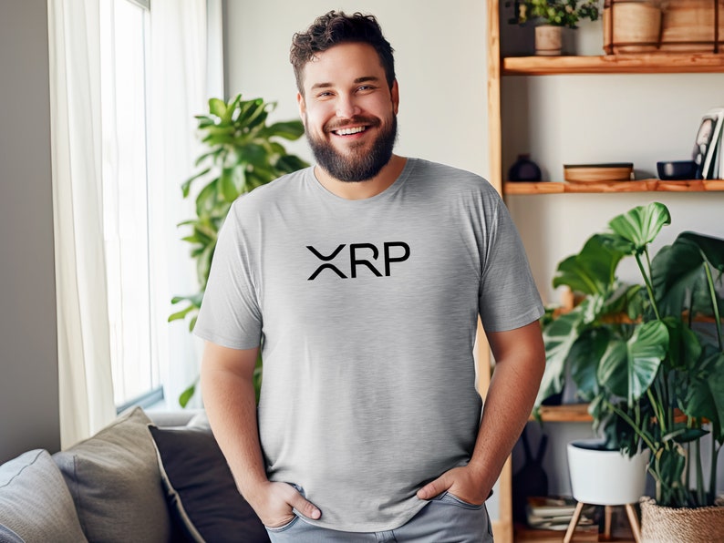 May include: A man wearing a gray t-shirt with the word "XRP" printed in black on the front. He is smiling and standing in a room with a window and plants.