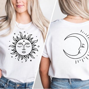 K&ouml;nnte beinhalten: Wei&szlig;es T-Shirt mit einem schwarzen Sonnen- und Monddesign. Die Sonne hat ein Gesicht mit geschlossenen Augen und der Mond hat ein Gesicht mit einem offenen Auge. Sonne und Mond sind von kleinen Sternen umgeben.
