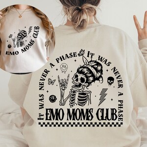 Puede incluir: Una sudadera beige con un estampado gr&aacute;fico negro de un esqueleto con una cabeza de calavera que lleva un mo&ntilde;o y hace un gesto de rock and roll. El texto alrededor del esqueleto dice "It was never a phase it was never a phase Emo Moms Club".