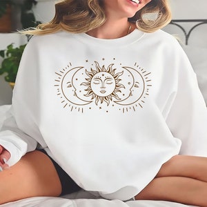 K&ouml;nnte beinhalten: Wei&szlig;er Pullover mit einem braunen Sonnen- und Mondmotiv. Sonne und Mond haben Gesichter und sind von Sternen umgeben.