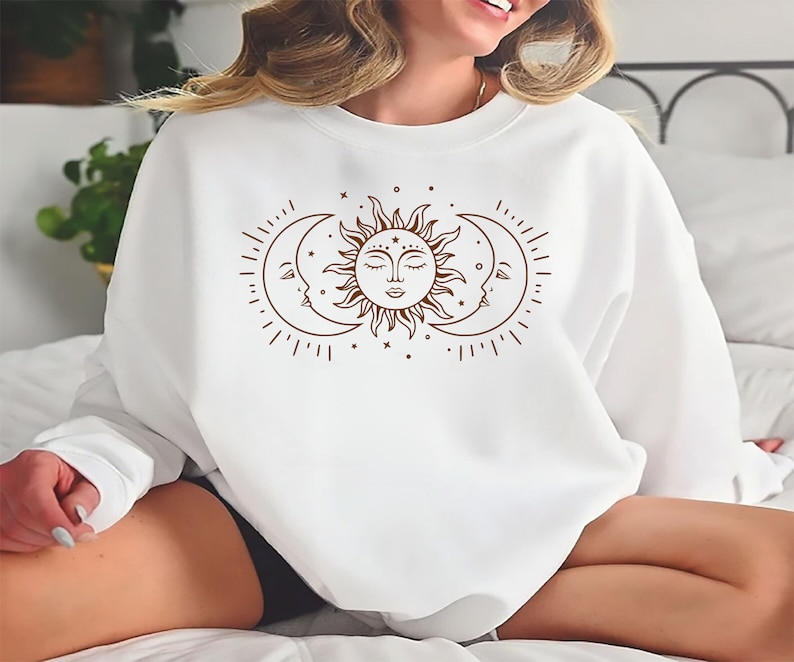 K&ouml;nnte beinhalten: Wei&szlig;er Pullover mit einem braunen Sonnen- und Mondmotiv. Sonne und Mond haben Gesichter und sind von Sternen umgeben.