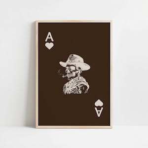 Op de afbeelding: Een bruine poster met een witte illustratie van een skelet dat een cowboyhoed draagt en een sigaret rookt. De poster heeft de tekst "A" en een hartje bovenaan en de tekst "A" en een hartje onderaan.