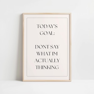 以下が含まれることがあります： 「TODAY'S GOAL: DONT SAY WHAT IM ACTUALLY THINKING」のテキストが黒いフォントでクリーム色の背景に印刷されたフレーム入りプリント。フレームは明るい木の色で、プリントは中央に配置されています。