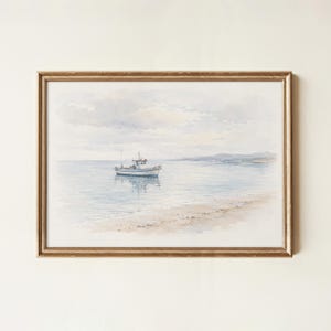 May include: A framed watercolor painting depicting a small white boat floating on calm blue water near a sandy shoreline under a soft, cloudy sky. The artwork is enclosed in a thin, metallic gold frame with a textured finish and neutral border.