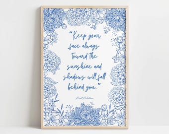 Op de afbeelding: Een ingelijst print met een blauw bloemenontwerp en een citaat van Walt Whitman: "Houd je gezicht altijd naar de zon gericht, en de schaduwen zullen achter je vallen."