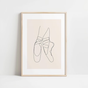 Op de afbeelding: Een minimalistische lijntekening in een lijst met een paar balletschoenen. Het kunstwerk is uitgevoerd in zwarte lijnen op een crèmekleurige achtergrond, in een licht houten frame. De afbeelding toont de elegantie van ballet.