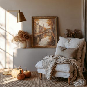 May include: A framed landscape painting depicting a canal-side village in autumn colors, displayed above a white armchair with a chunky knit throw blanket, a brass floor lamp, a vase of dried flowers, and three orange pumpkins on the floor.