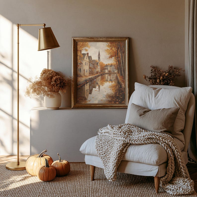 May include: A framed landscape painting depicting a canal-side village in autumn colors, displayed above a white armchair with a chunky knit throw blanket, a brass floor lamp, a vase of dried flowers, and three orange pumpkins on the floor.