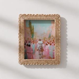 May include: A framed print depicting a religious scene with a figure on a donkey, surrounded by a crowd. The artwork features pastel colors and is set within a woven, rectangular frame. The frame is made of a light brown material.